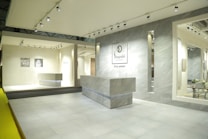 An interior space featuring a modern showroom with large ceramic tiles and bright lighting. The walls display brand signage and various tile samples. A sleek, minimalist desk is centered, surrounded by a clean and spacious environment.