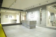 An interior space featuring a modern showroom with large ceramic tiles and bright lighting. The walls display brand signage and various tile samples. A sleek, minimalist desk is centered, surrounded by a clean and spacious environment.