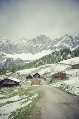A rustic mountain village scene with several wooden cabins nestled in a lush, green landscape with patches of snow. The foreground features a dirt path leading up to the cabins, while majestic, snow-capped mountains rise in the background.