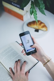 A person holds a smartphone in one hand while the other hand is on an open book. The smartphone screen displays a login interface. Nearby, a croissant rests on a white plate. Overhead, a plant with green leaves hangs over the table.