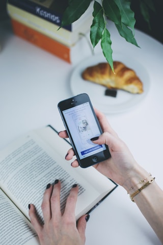 A person holds a smartphone in one hand while the other hand is on an open book. The smartphone screen displays a login interface. Nearby, a croissant rests on a white plate. Overhead, a plant with green leaves hangs over the table.