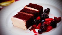 A slice of rich chocolate cake with creamy white frosting is plated alongside a variety of fresh berries, including blackberries, blueberries, and sliced strawberries, all set on a white dish.