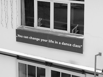 A sign with the motivational quote 'You can change your life in a dance class' is affixed below a window on the exterior of a building. The sign is placed on a ledge covered with a thin layer of snow, and there are decorative lights hanging nearby.