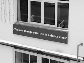 A sign with the motivational quote 'You can change your life in a dance class' is affixed below a window on the exterior of a building. The sign is placed on a ledge covered with a thin layer of snow, and there are decorative lights hanging nearby.