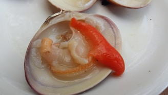 An open clam shell containing a portion of seafood and a reddish-orange piece, possibly a type of pepper or garnish, placed on a white surface.