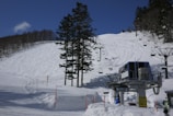 A mountainous ski slope covered in fresh snow, marked by ski tracks. A ski lift with chairs is visible, stretching up the slope. A control station or platform for the lift is positioned at the base. Tall evergreen trees partially obscure the view of the slope, and a clear blue sky with a few thin clouds enhances the winter landscape.