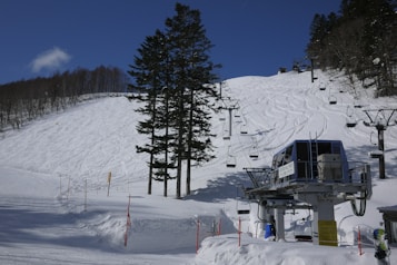A mountainous ski slope covered in fresh snow, marked by ski tracks. A ski lift with chairs is visible, stretching up the slope. A control station or platform for the lift is positioned at the base. Tall evergreen trees partially obscure the view of the slope, and a clear blue sky with a few thin clouds enhances the winter landscape.