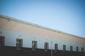 A building with a light blue facade and multiple windows under a clear blue sky. The roof is corrugated and the walls are made of a dark material with a rough texture at the bottom and smooth above.