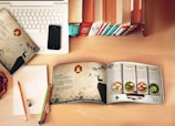 A workspace setup with a laptop partially covered by a patterned folder featuring a deer and nature scene. An open booklet displays recipes with colorful images of food dishes on a wooden table. A smartphone rests on the laptop, and a row of upright books adds a cozy, library-like feel. Colorful pencils rest on a notepad with some geometric sketches and an eraser nearby on a wooden desk.