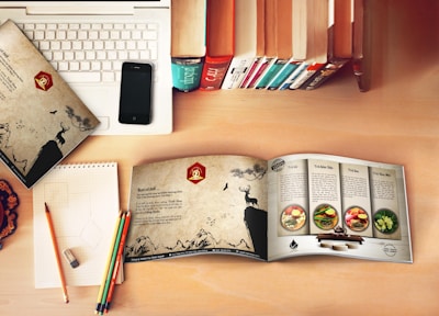 A workspace setup with a laptop partially covered by a patterned folder featuring a deer and nature scene. An open booklet displays recipes with colorful images of food dishes on a wooden table. A smartphone rests on the laptop, and a row of upright books adds a cozy, library-like feel. Colorful pencils rest on a notepad with some geometric sketches and an eraser nearby on a wooden desk.