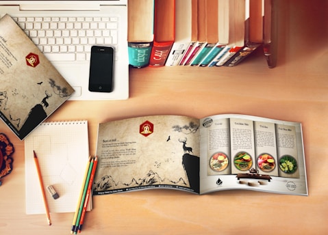 A workspace setup with a laptop partially covered by a patterned folder featuring a deer and nature scene. An open booklet displays recipes with colorful images of food dishes on a wooden table. A smartphone rests on the laptop, and a row of upright books adds a cozy, library-like feel. Colorful pencils rest on a notepad with some geometric sketches and an eraser nearby on a wooden desk.