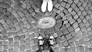 Two pairs of feet facing each other on a cobblestone pavement. One person is wearing white shoes while the other is wearing black sneakers with white laces. They are standing around an object embedded in the ground that appears to be a circular metal plate.