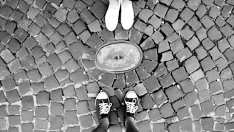 Two pairs of feet facing each other on a cobblestone pavement. One person is wearing white shoes while the other is wearing black sneakers with white laces. They are standing around an object embedded in the ground that appears to be a circular metal plate.