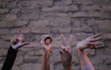 Several hands are forming the word 'LOVE' using sign language against a textured stone wall. Each hand is positioned to represent a different letter, with various wrist accessories visible.