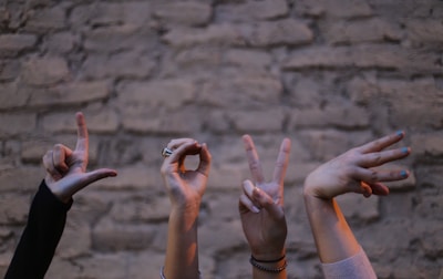 Several hands are forming the word 'LOVE' using sign language against a textured stone wall. Each hand is positioned to represent a different letter, with various wrist accessories visible.