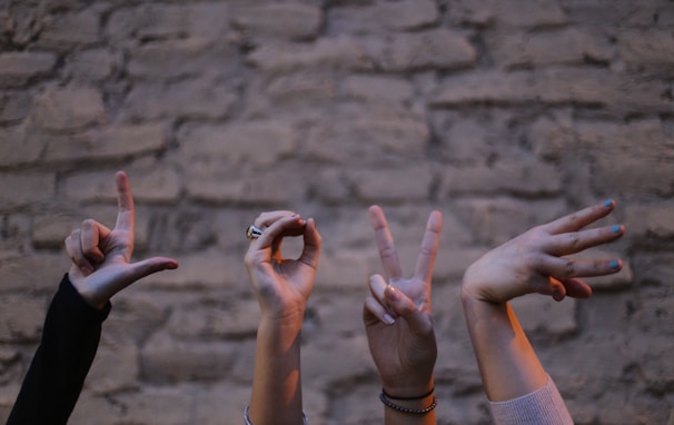 Several hands are forming the word 'LOVE' using sign language against a textured stone wall. Each hand is positioned to represent a different letter, with various wrist accessories visible.