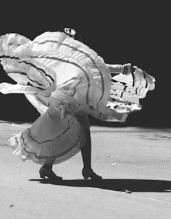 A black and white image of a dancer captured in mid-movement, showcasing the flowing, ruffled layers of a traditional dress. The play of light and shadow emphasizes the motion and texture of the fabric.