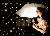A person holding a white umbrella is seated in a dark, possibly forested, environment. They are wearing a plaid shirt and looking upward, surrounded by numerous glowing orbs resembling fireflies or stars, creating a magical and serene atmosphere.