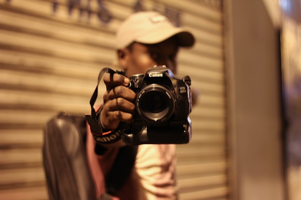 A person wearing a hat is holding a Canon EOS 60D camera, focusing towards the viewer. The background is a blurred metallic shutter, suggesting an urban setting. The atmosphere is dimly lit, likely due to evening or indoor lighting.