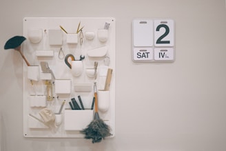 A beige wall features an organized pegboard with multiple compartments and hooks holding various office supplies and cleaning tools, such as rulers, brushes, scissors, and small containers. To the right, a minimalistic wall calendar displays the date as 'SAT 2 APRIL.'