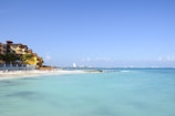A tranquil beach scene featuring a sandy shoreline with several lounge chairs and umbrellas, adjacent to a resort with colorful buildings. The calm, turquoise ocean extends to the horizon under a clear blue sky. In the distance, a city skyline is faintly visible.