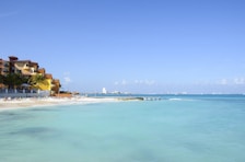 A tranquil beach scene featuring a sandy shoreline with several lounge chairs and umbrellas, adjacent to a resort with colorful buildings. The calm, turquoise ocean extends to the horizon under a clear blue sky. In the distance, a city skyline is faintly visible.