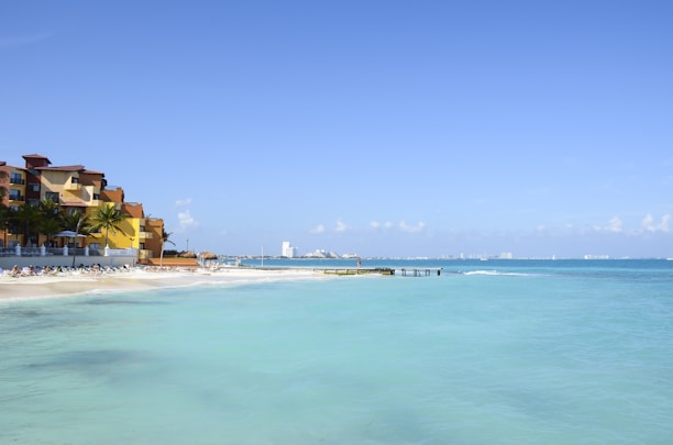 A tranquil beach scene featuring a sandy shoreline with several lounge chairs and umbrellas, adjacent to a resort with colorful buildings. The calm, turquoise ocean extends to the horizon under a clear blue sky. In the distance, a city skyline is faintly visible.