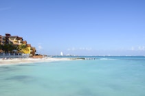 A tranquil beach scene featuring a sandy shoreline with several lounge chairs and umbrellas, adjacent to a resort with colorful buildings. The calm, turquoise ocean extends to the horizon under a clear blue sky. In the distance, a city skyline is faintly visible.