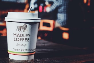 A disposable coffee cup with a logo and text sits on a wooden table. The background is blurred, featuring a person in a blue checkered shirt sitting by a window.
