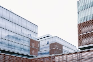 Modern architectural structures with glass facades and horizontal wooden slats are featured against a pale sky. The buildings display clean lines and a minimalist design, emphasizing transparency and simplicity.