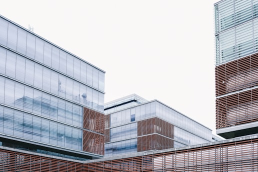 Modern architectural structures with glass facades and horizontal wooden slats are featured against a pale sky. The buildings display clean lines and a minimalist design, emphasizing transparency and simplicity.