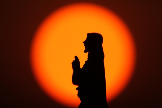 A silhouette of a person with clasped hands, possibly in prayer, against a vibrant, large orange sunset. The background is an intense, glowing gradient of orange and red, creating a dramatic contrast with the black silhouette.