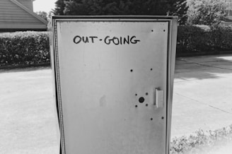 A black and white photograph of a metal utility box with the words 'OUT-GOING' written on it. The box has several small holes and grommet-like features. It is located on a sidewalk with hedges and trees visible in the background, and a house partially visible on one side.