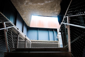 A metal staircase leads upward, with railings on both sides made of metal mesh. The scene is framed by industrial architecture, with concrete and steel beams. Natural light enters from an opening above, casting shadows on the steps.