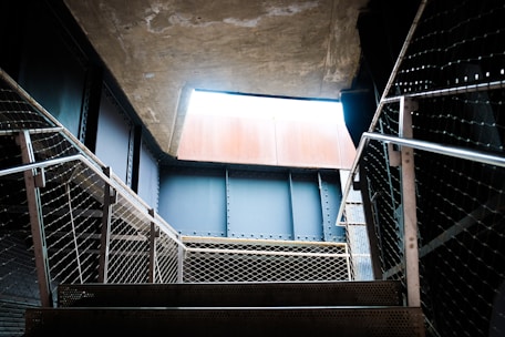 A metal staircase leads upward, with railings on both sides made of metal mesh. The scene is framed by industrial architecture, with concrete and steel beams. Natural light enters from an opening above, casting shadows on the steps.