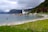 A serene coastal scene featuring a row of small houses along the shoreline with a white church in the background. The landscape includes lush green grass, a sandy beach, and calm waters. Hills rise prominently in the background under a cloudy sky.
