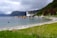 A serene coastal scene featuring a row of small houses along the shoreline with a white church in the background. The landscape includes lush green grass, a sandy beach, and calm waters. Hills rise prominently in the background under a cloudy sky.