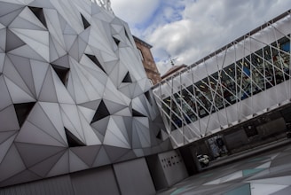 A modern architectural structure featuring a geometric façade made up of triangular patterns and a skybridge with a lattice design. The building displays a combination of contemporary design elements with a focus on angular shapes. There is a contrast between the gray, metallic surface and the colorful stained glass-like elements visible in the bridge section.