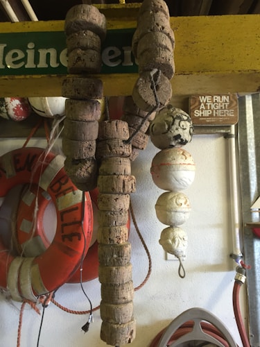 A collection of nautical items displayed on a wall includes multiple cork floats, life preservers, and a wooden sign with text. The scene has a rustic and nautical theme, with visible textures of cork and rope. A green and yellow branded sign is partially visible at the top. There is also a small amount of visible wiring.