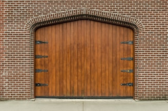 A large, arched wooden door set within a brick wall, featuring decorative black metal hinges on its surface. The bricks are arranged in a symmetrical pattern, creating a classic architectural style.