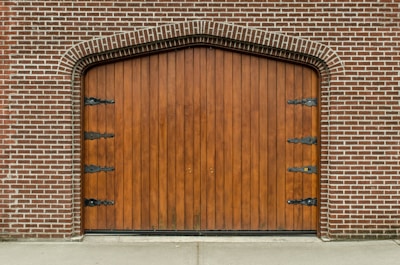 A large, arched wooden door set within a brick wall, featuring decorative black metal hinges on its surface. The bricks are arranged in a symmetrical pattern, creating a classic architectural style.
