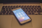 A smartphone displaying a call screen rests on the keyboard of a laptop. The laptop keyboard is slightly out of focus, and the smartphone shows an ongoing call with several icons for call options.