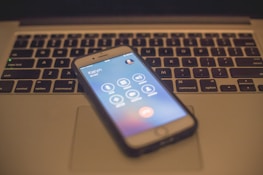 A smartphone displaying a call screen rests on the keyboard of a laptop. The laptop keyboard is slightly out of focus, and the smartphone shows an ongoing call with several icons for call options.