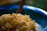 A close-up of sugar-coated ginger pieces piled in a blue mixing bowl. The ginger pieces are cubed and have a sugary texture, with a wooden spoon partially visible in the background. The focus is on the ginger, while the bowl and background are softly blurred.