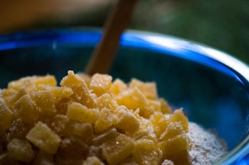 A close-up of sugar-coated ginger pieces piled in a blue mixing bowl. The ginger pieces are cubed and have a sugary texture, with a wooden spoon partially visible in the background. The focus is on the ginger, while the bowl and background are softly blurred.
