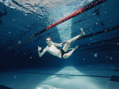 A person swimming underwater with goggles and giving a thumbs-up, surrounded by the clear blue water of a swimming pool. The serene environment is enhanced by the light patterns on the pool floor and the colorful lane divider above.