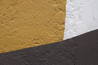 A textured wall surface featuring three distinct sections painted in mustard yellow, white, and dark gray tones. Each color has a rough, uneven texture that suggests the wall is made of plaster or concrete.