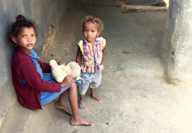 Two young children are in a dimly lit, bare room. One child, sitting on the ground, is holding a teddy bear and wearing a red sweater and blue dress. The other child stands next to the seated child, wearing a striped vest and shorts. The walls and floor are concrete, giving a stark and minimal appearance.