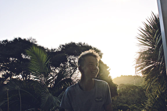 A warm photo of Kandau smiling outdoors, surrounded by lush greenery and sunlight filtering through trees.