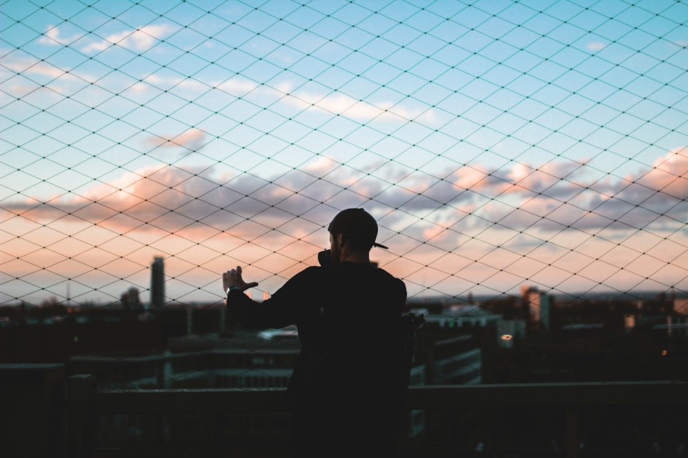 man standing behind chain link fence holding to fence during golden hour